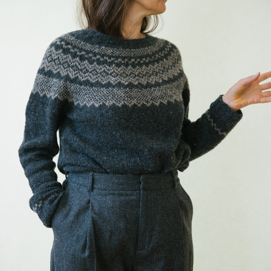 A woman wearing a dark grey handknitted sweater and seemingly talking, seen from the front