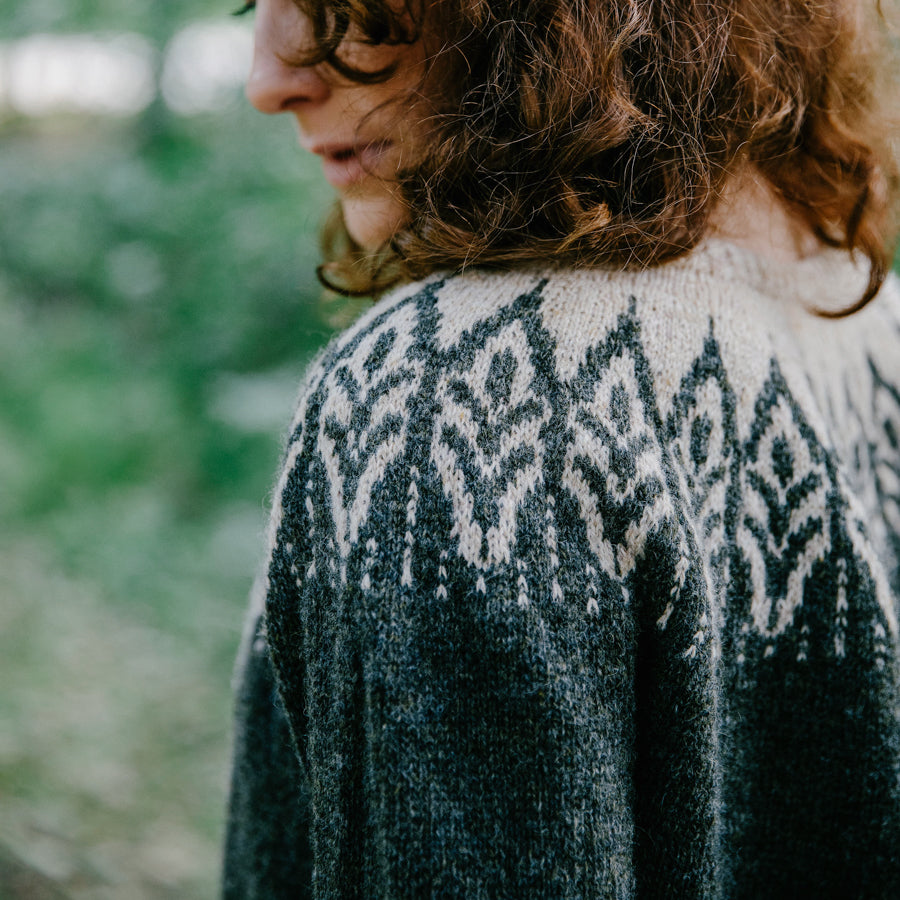 A young woman in the forest wearing a dark green handknitted sweater, seen from the side/back