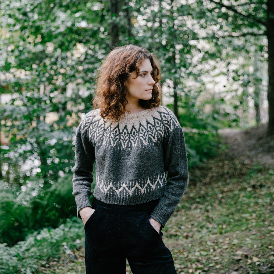 A young woman in the forest wearing a dark green handknitted sweater, seen from the front