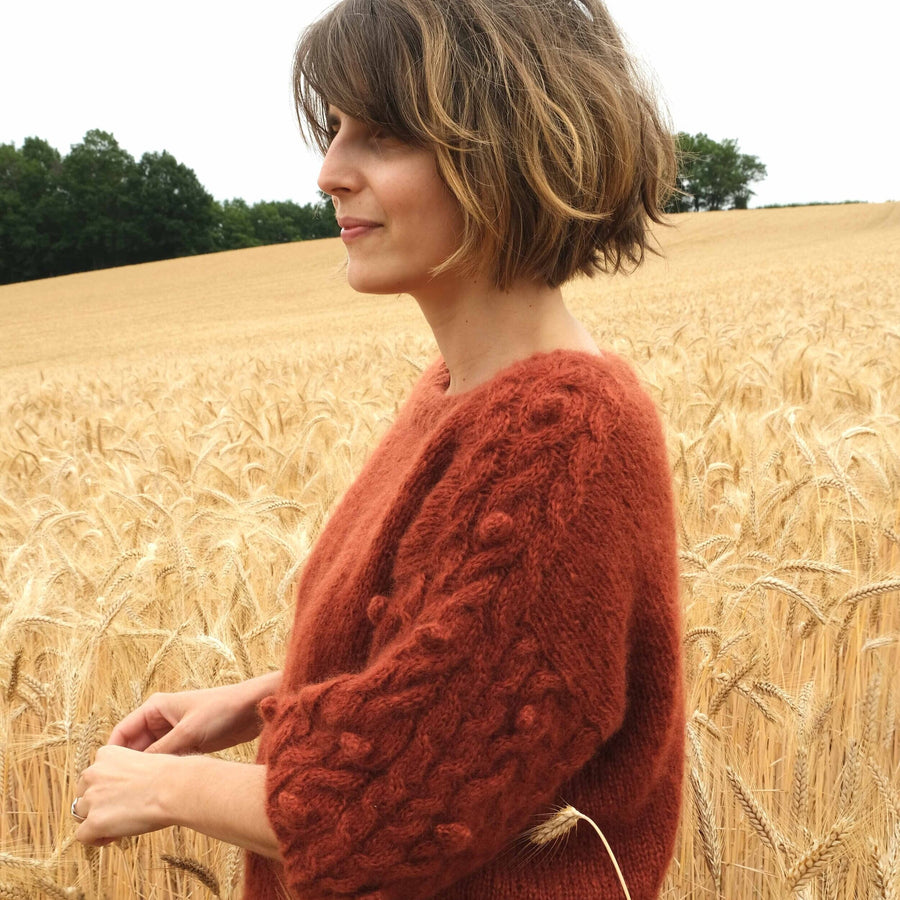 a young woman wearing a red handknitted sweater in a corn field, seen from the side