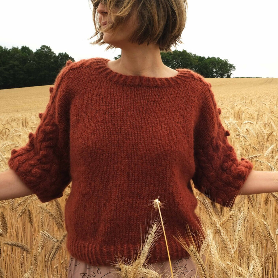 a young woman wearing a red handknitted sweater in a corn field, seen from the front