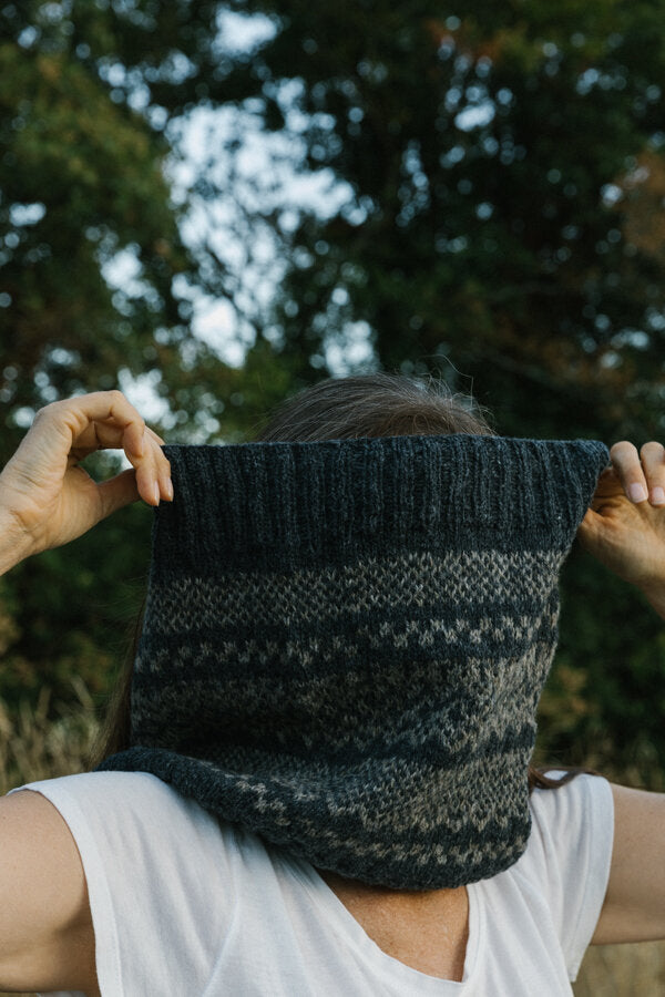 A woman wearing a handknitted Afterparty cowl in front of her face, shown in nature